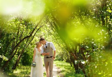 A bride and groom walking hand in hand through a lush park path, both lean into one another with their foreheads touching.
