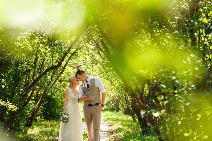 A bride and groom walking hand in hand through a lush park path, both lean into one another with their foreheads touching.