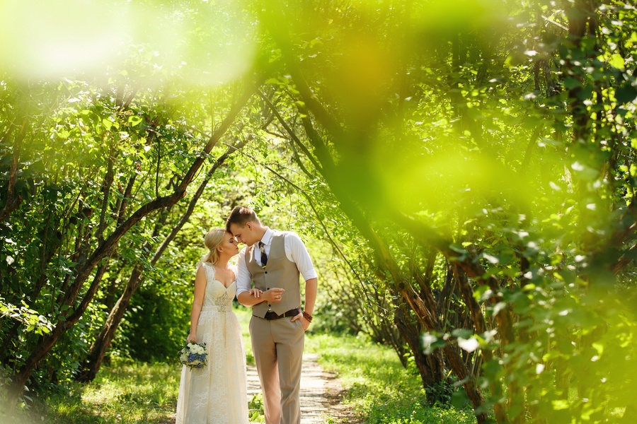 A bride and groom walking hand in hand through a lush park path, both lean into one another with their foreheads touching.