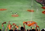 Fans in stadium stands wave orange towels while a baseball game unfolds below with players gathered on the pitcher’s mound.