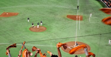 Fans in stadium stands wave orange towels while a baseball game unfolds below with players gathered on the pitcher’s mound.