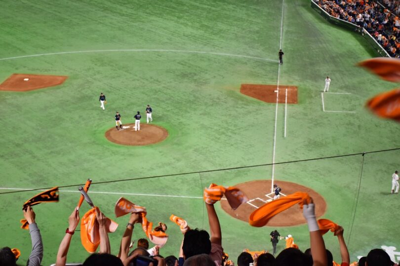 Fans in stadium stands wave orange towels while a baseball game unfolds below with players gathered on the pitcher’s mound.