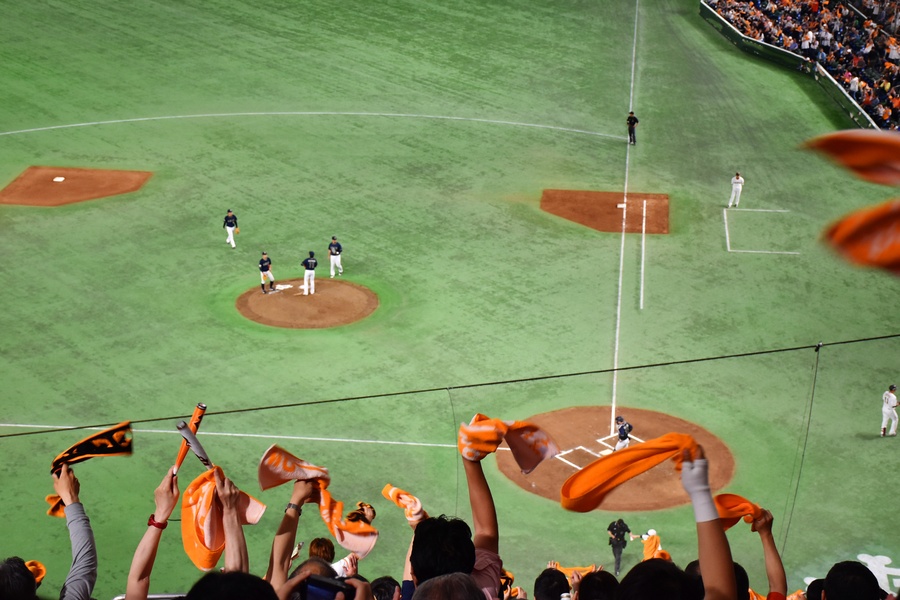Fans in stadium stands wave orange towels while a baseball game unfolds below with players gathered on the pitcher’s mound.