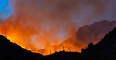 A mountain range is in the background and in the foreground there is a fire blazing. Smoke is rising from the fire.