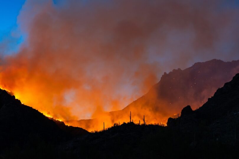 A mountain range is in the background and in the foreground there is a fire blazing. Smoke is rising from the fire.