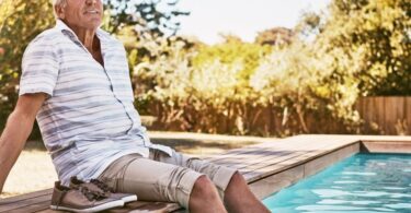 A close-up of an older man sitting by a swimming pool with his feet in the water. His shoes are on the deck next to him.