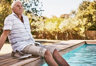 A close-up of an older man sitting by a swimming pool with his feet in the water. His shoes are on the deck next to him.