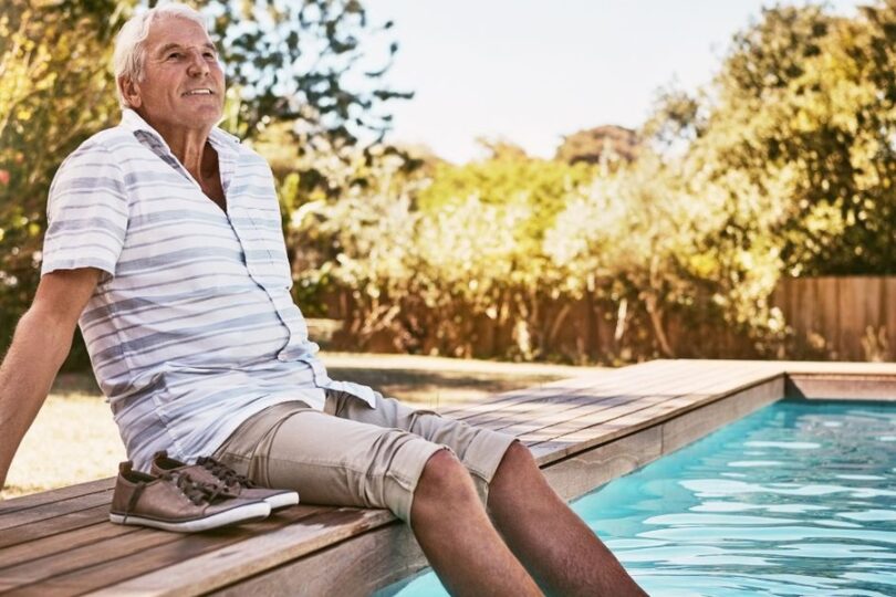 A close-up of an older man sitting by a swimming pool with his feet in the water. His shoes are on the deck next to him.