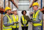 A group of warehouse workers is having a meeting in an aisle of industrial shelves. One of them is holding a laptop.