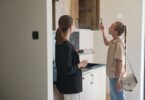 Woman opens an upper kitchen cabinet while another woman with a clipboard observes in a bright apartment.