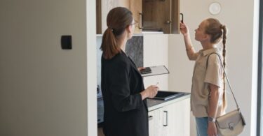Woman opens an upper kitchen cabinet while another woman with a clipboard observes in a bright apartment.