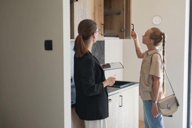 Woman opens an upper kitchen cabinet while another woman with a clipboard observes in a bright apartment.
