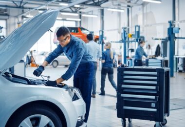 A man wears safety goggles and gloves while working on a car engine with a wrench in a brightly lit body shop.