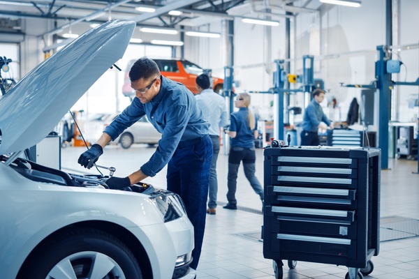 A man wears safety goggles and gloves while working on a car engine with a wrench in a brightly lit body shop.