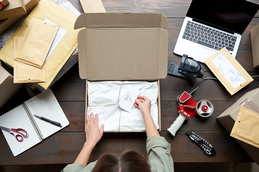 Someone stands at a desk with packaging supplies. They have an open product box with twine and a "thank you" card inside.