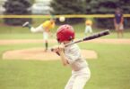 A small child wearing a baseball uniform and helmet holds a bat above his shoulder. Other kids are blurred in the outfield.