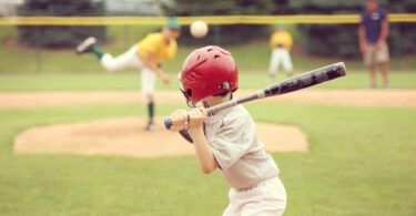 A small child wearing a baseball uniform and helmet holds a bat above his shoulder. Other kids are blurred in the outfield.