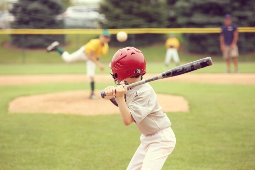 A small child wearing a baseball uniform and helmet holds a bat above his shoulder. Other kids are blurred in the outfield.