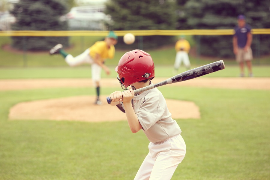 A small child wearing a baseball uniform and helmet holds a bat above his shoulder. Other kids are blurred in the outfield.