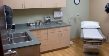 A clean medical exam room with ran cabinets and a grey countertop. A sheeted exam table sits in the corner of the room.