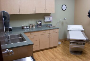 A clean medical exam room with ran cabinets and a grey countertop. A sheeted exam table sits in the corner of the room.