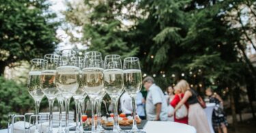 Several glasses of champagne sit on a table at an outdoor gala. Several people stand in the background.