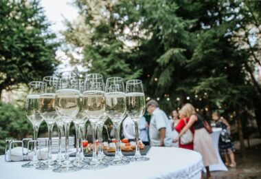 Several glasses of champagne sit on a table at an outdoor gala. Several people stand in the background.