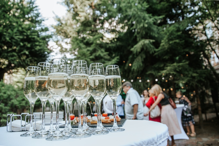 Several glasses of champagne sit on a table at an outdoor gala. Several people stand in the background.
