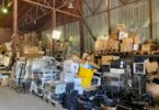  Warehouse filled with stacked boxes, pallets, wrapped equipment, loose monitors, and electronics under metal rafters.