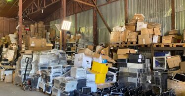  Warehouse filled with stacked boxes, pallets, wrapped equipment, loose monitors, and electronics under metal rafters.