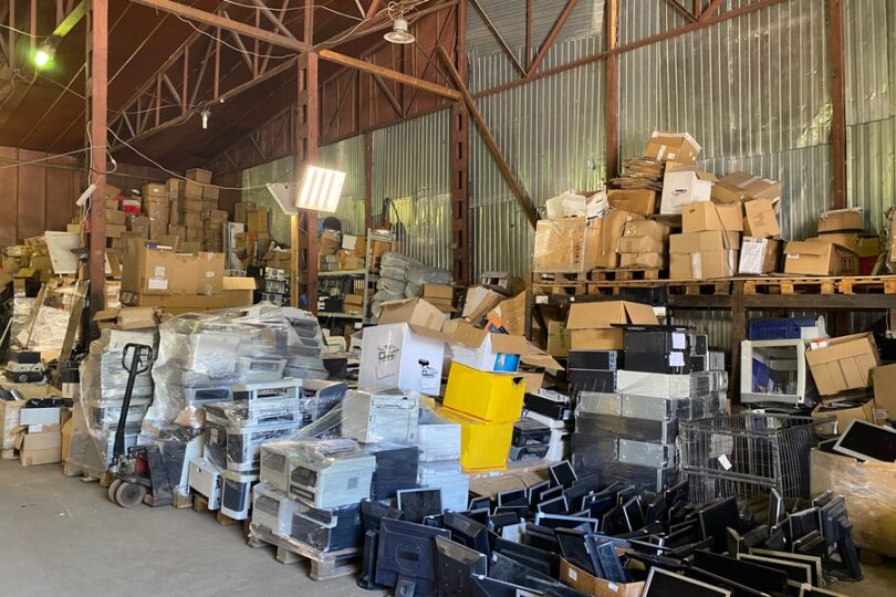  Warehouse filled with stacked boxes, pallets, wrapped equipment, loose monitors, and electronics under metal rafters.