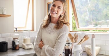 A smiling woman standing in the kitchen of her home. There are appliances on the counter and the window is open.