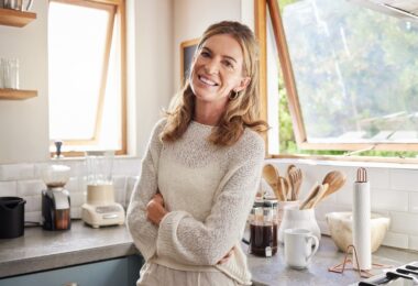 A smiling woman standing in the kitchen of her home. There are appliances on the counter and the window is open.