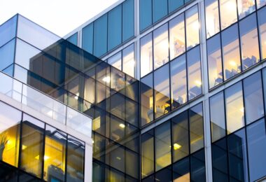 Close-up of a contemporary office exterior with blue-tinted glass, warm lights, and workstations visible inside.