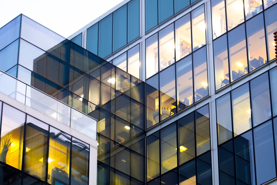 Close-up of a contemporary office exterior with blue-tinted glass, warm lights, and workstations visible inside.