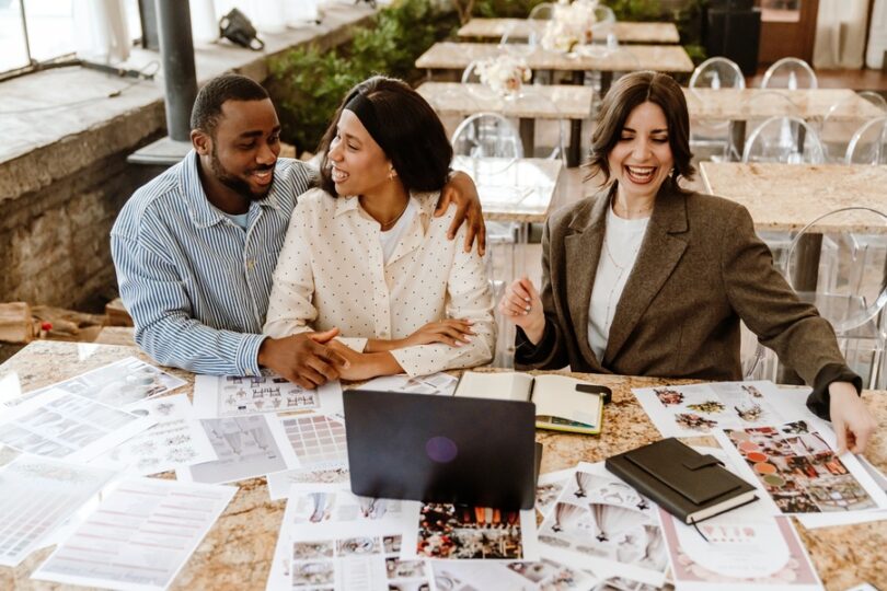 A wedding consultant and a couple sitting with a laptop. There are printed planning sheets covering the table.