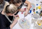 Two women in an art studio shaping small bits of clay. One of the women is reaching over to help the other one.