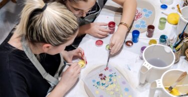 Two women in an art studio shaping small bits of clay. One of the women is reaching over to help the other one.