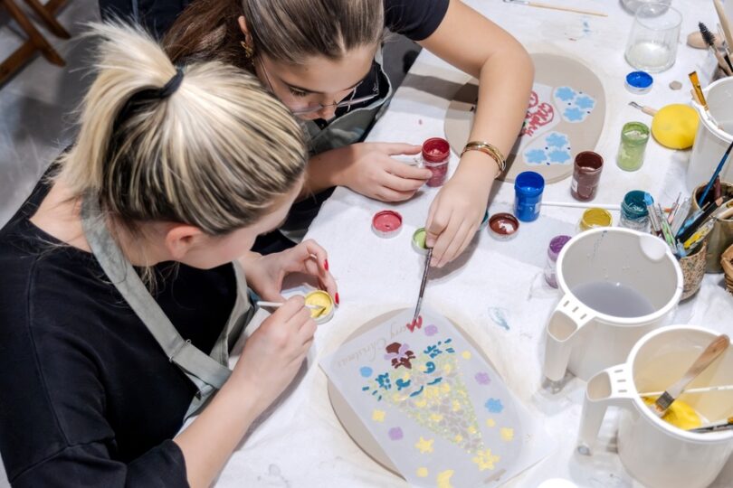 Two women in an art studio shaping small bits of clay. One of the women is reaching over to help the other one.