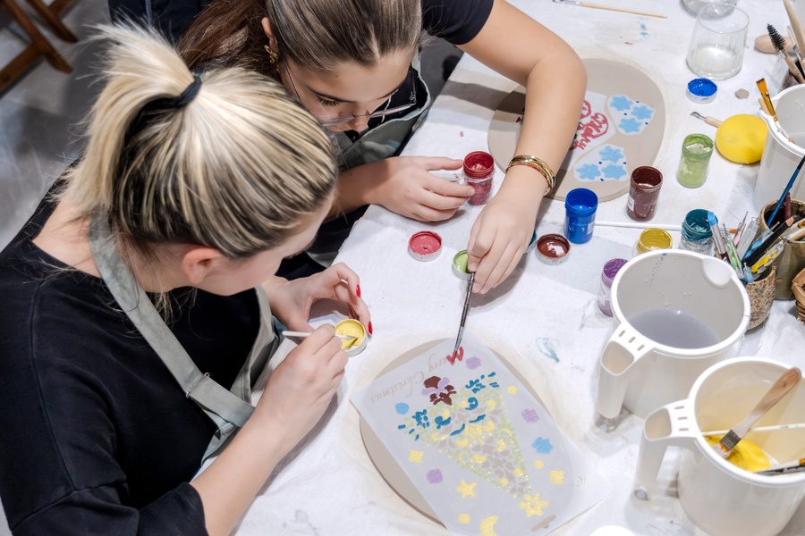 Two women in an art studio shaping small bits of clay. One of the women is reaching over to help the other one.