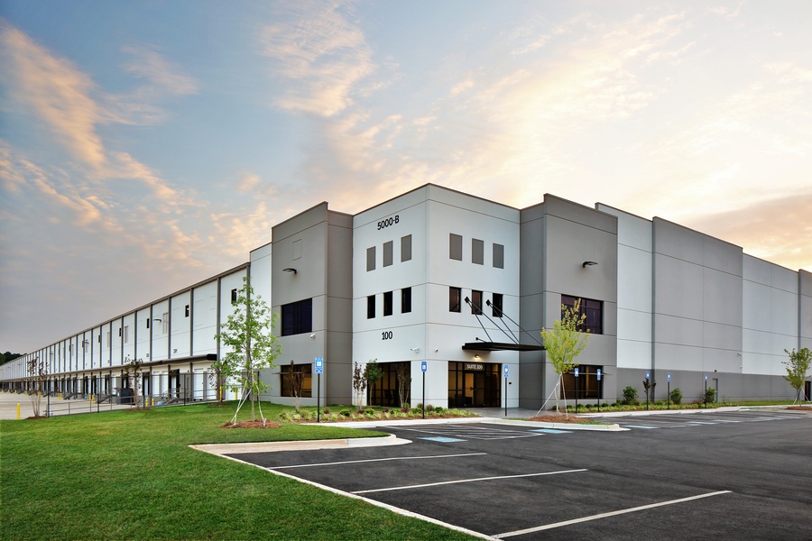 A white-and-tan commercial warehouse features a large parking lot. The sun is setting behind the facility.
