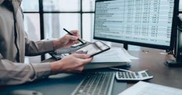 A person sits at their desk, looking at their tablet while examining financials on their computer screen.