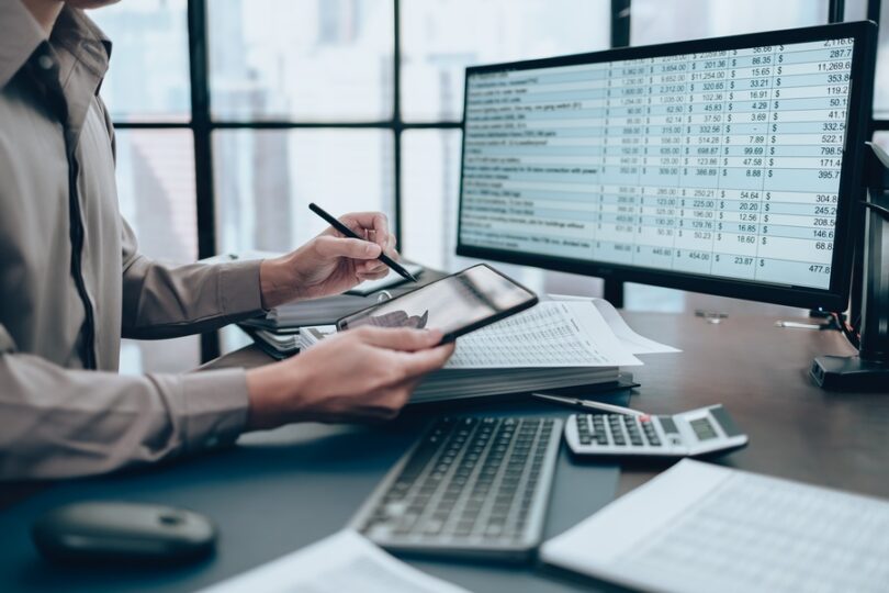 A person sits at their desk, looking at their tablet while examining financials on their computer screen.