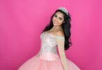 A young woman wearing a jeweled tiara and pink embellished dress while smiling against a bright pink background.