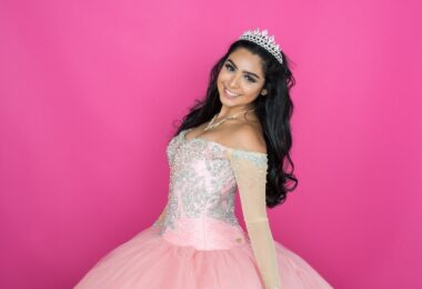 A young woman wearing a jeweled tiara and pink embellished dress while smiling against a bright pink background.