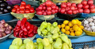a farmer's market display with tomatoes, peppers, eggplants, cucumbers, lemons, onions, and garlic in baskets.