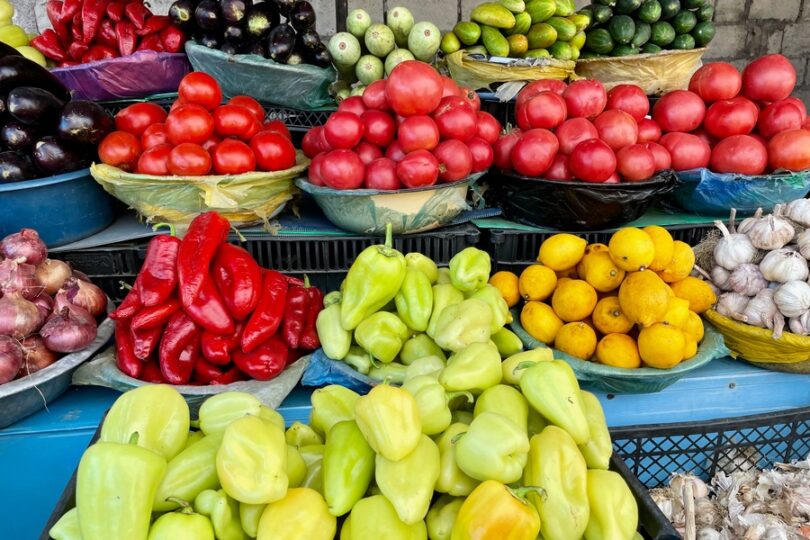 a farmer's market display with tomatoes, peppers, eggplants, cucumbers, lemons, onions, and garlic in baskets.