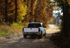A white pickup truck driving down a dirt road during the daytime. The road is surrounded by tall trees.