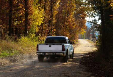 A white pickup truck driving down a dirt road during the daytime. The road is surrounded by tall trees.