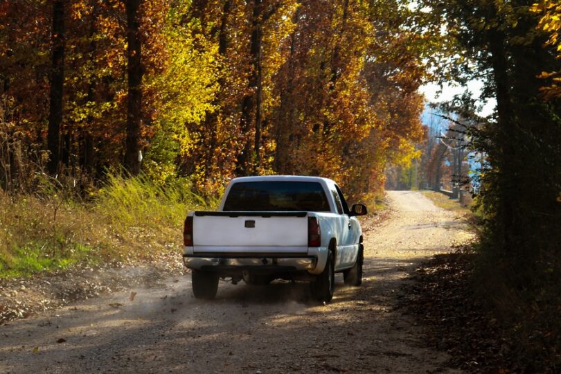 A white pickup truck driving down a dirt road during the daytime. The road is surrounded by tall trees.